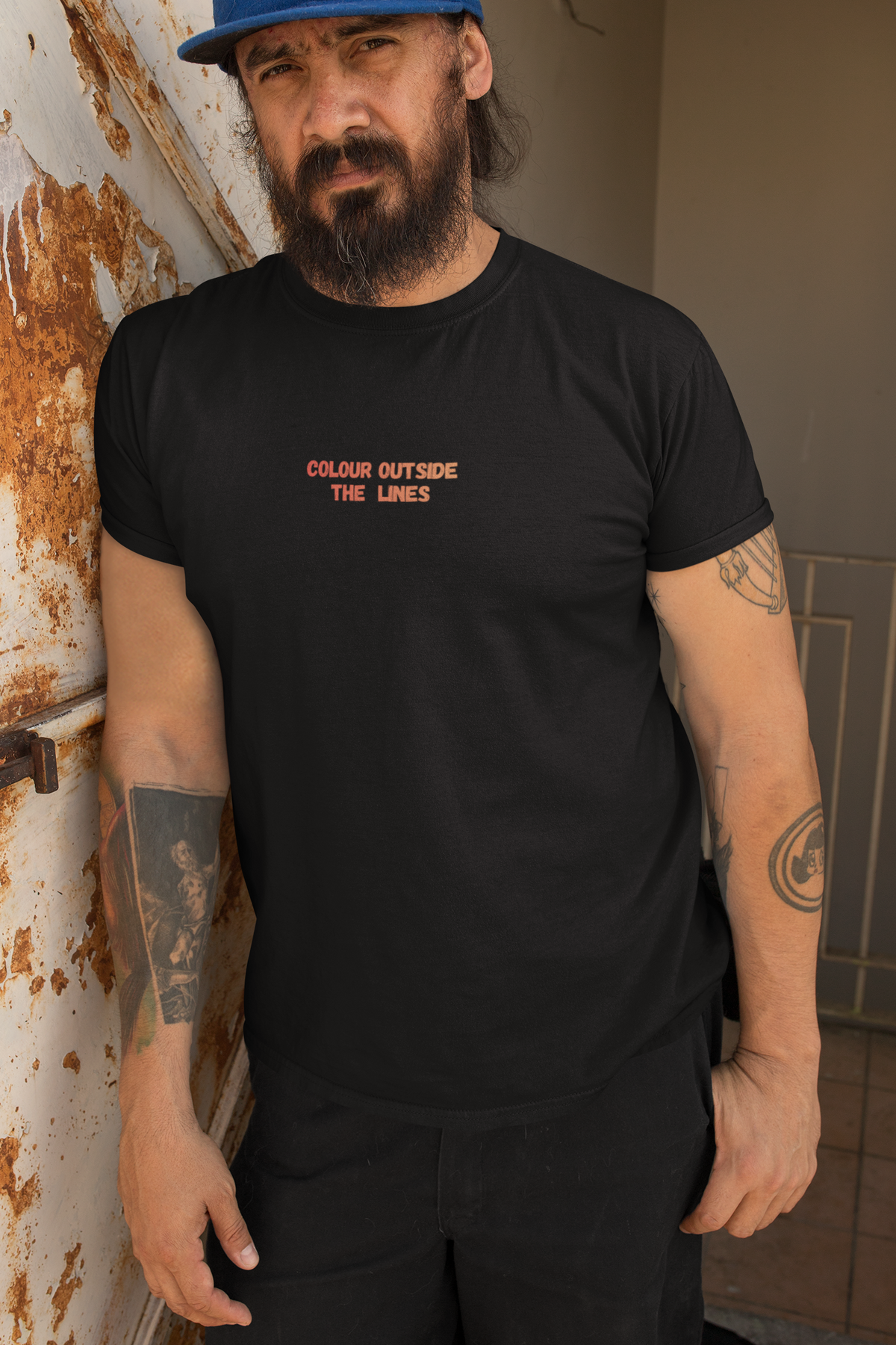 A man wearing a black crewneck t-shirt with the text 'COLOUR OUTSIDE THE LINES' printed in orange on the chest, standing in front of a rusted metal door.