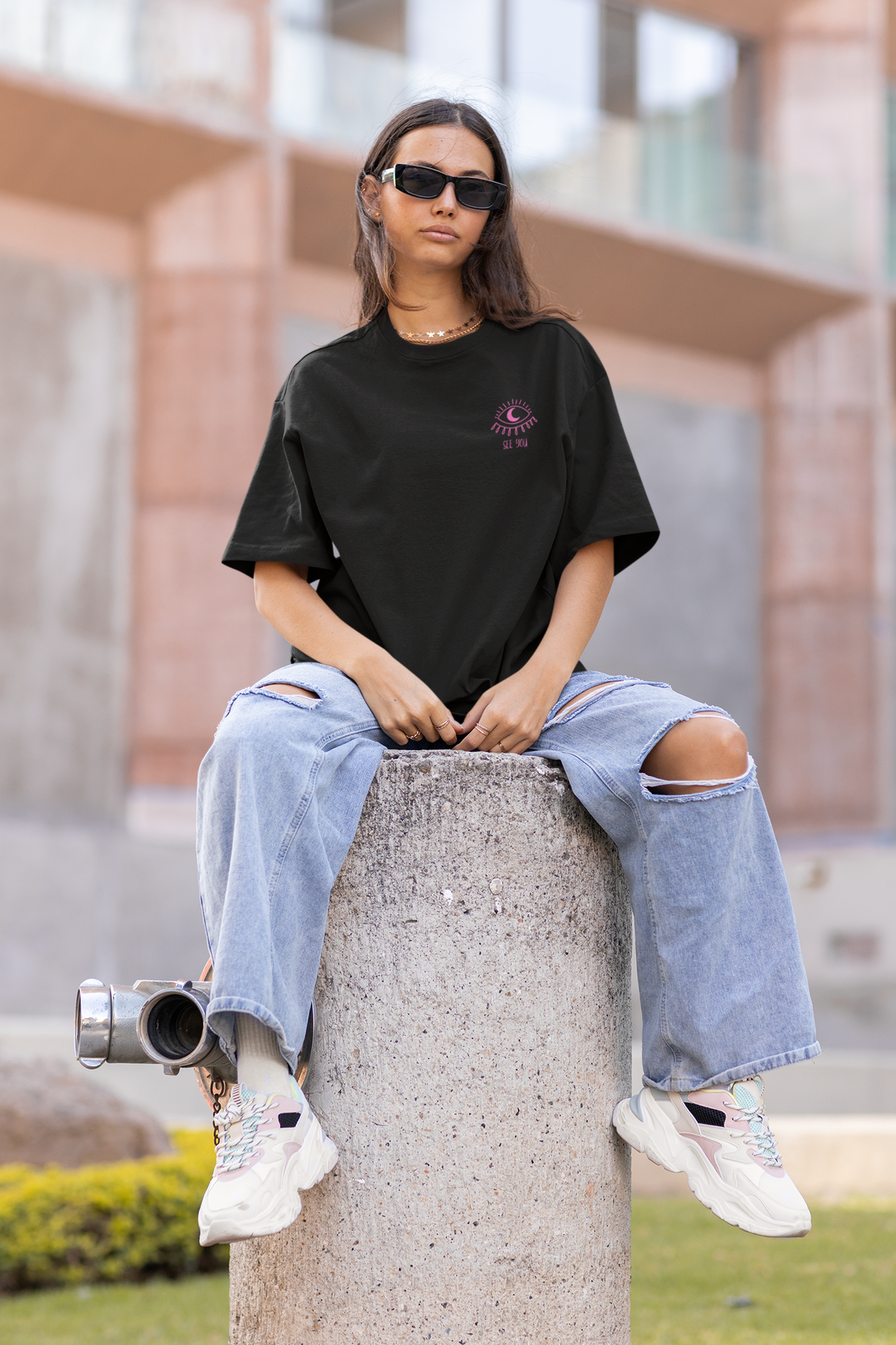 A woman sits on a concrete pillar outdoors wearing an oversized black t-shirt with a small purple graphic on the left chest, light-wash ripped jeans, white sneakers, and black sunglasses.