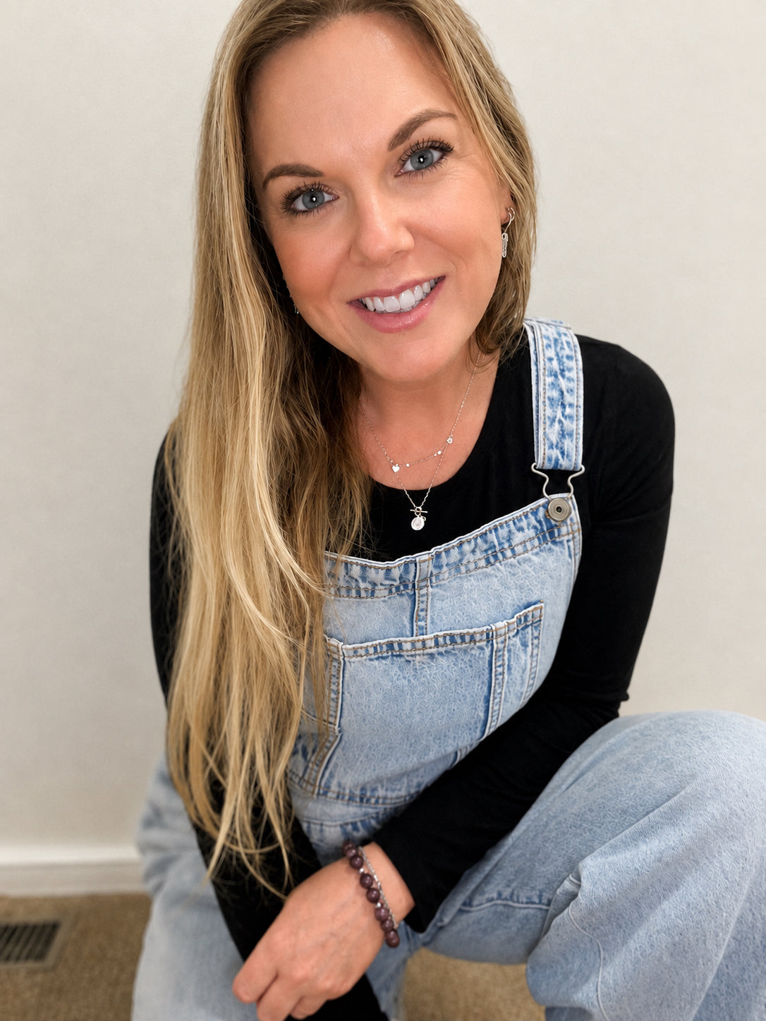 A smiling blonde woman wearing light-wash denim overalls over a black long-sleeve top, posing indoors against a plain light wall.