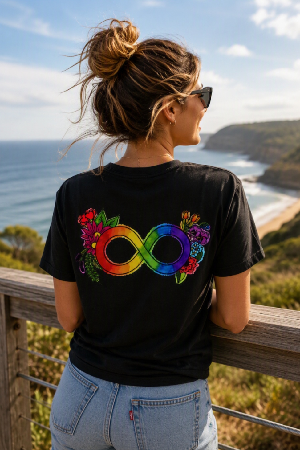 A woman wearing a black t-shirt with a colorful rainbow infinity symbol and floral designs on the back, standing outdoors by a wooden railing with a scenic ocean view.