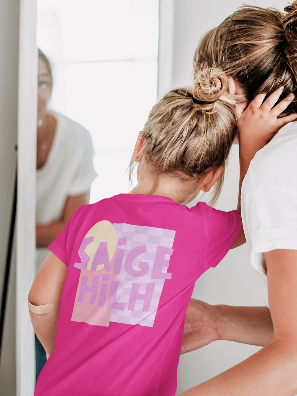 A young girl seen from behind wearing a bright pink t-shirt with a 'SAIGE HILH' graphic on the back, standing next to an adult woman in a white t-shirt in a brightly lit indoor setting.