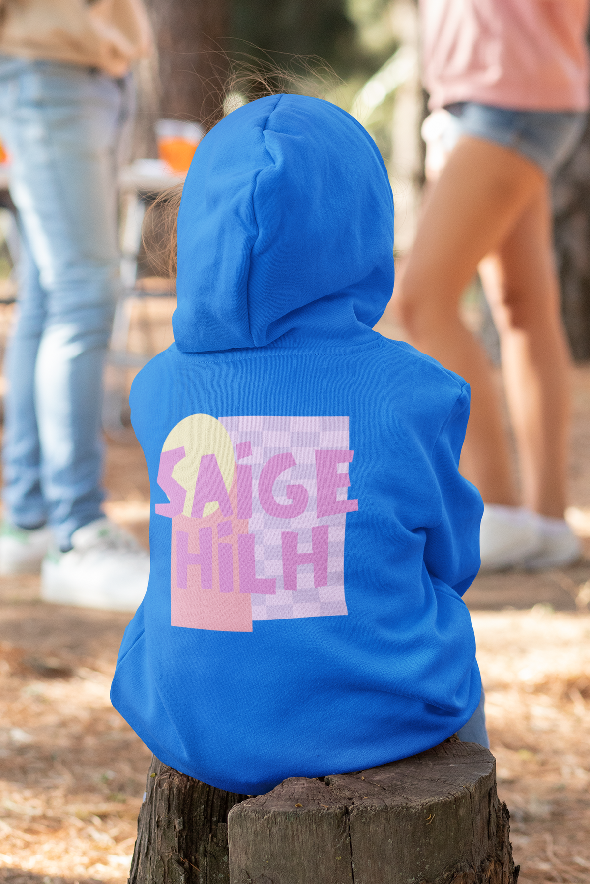 A child wearing a bright blue hoodie sits on a tree stump outdoors, viewed from behind. The hoodie has a large graphic on the back with the text 'SAIGE HILH' in pink over a pastel geometric design.