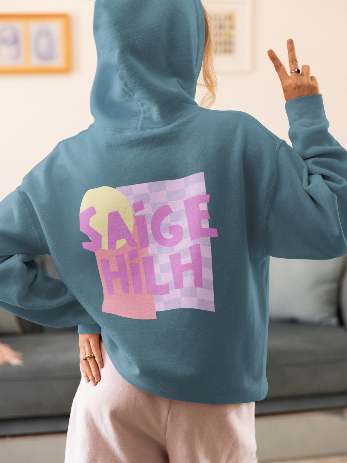 Person wearing a dusty blue hoodie featuring a large pastel pink, purple and yellow "Saige Hilh" graphic design printed on the back, shown indoors from the rear.