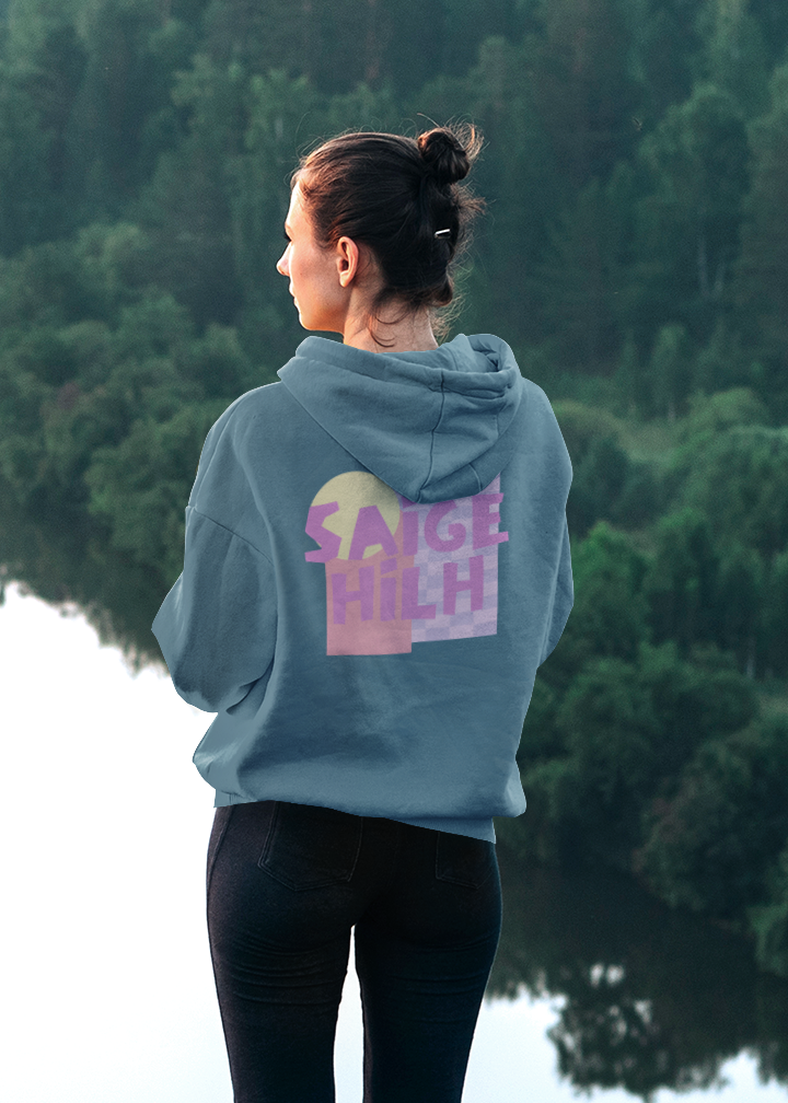 Person wearing a dusty blue hoodie featuring a large pastel pink, purple and yellow "Saige Hilh" graphic design printed on the back, shown indoors from the rear.