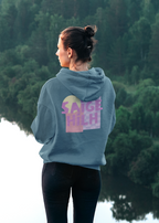 Person wearing a dusty blue hoodie featuring a large pastel pink, purple and yellow "Saige Hilh" graphic design printed on the back, shown indoors from the rear.