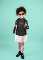 A young girl with wearing a black graphic hoodie, black leather jacket, a light pink tulle skirt, pink heart-patterned tights, and black boots, posing against a solid mint green background.