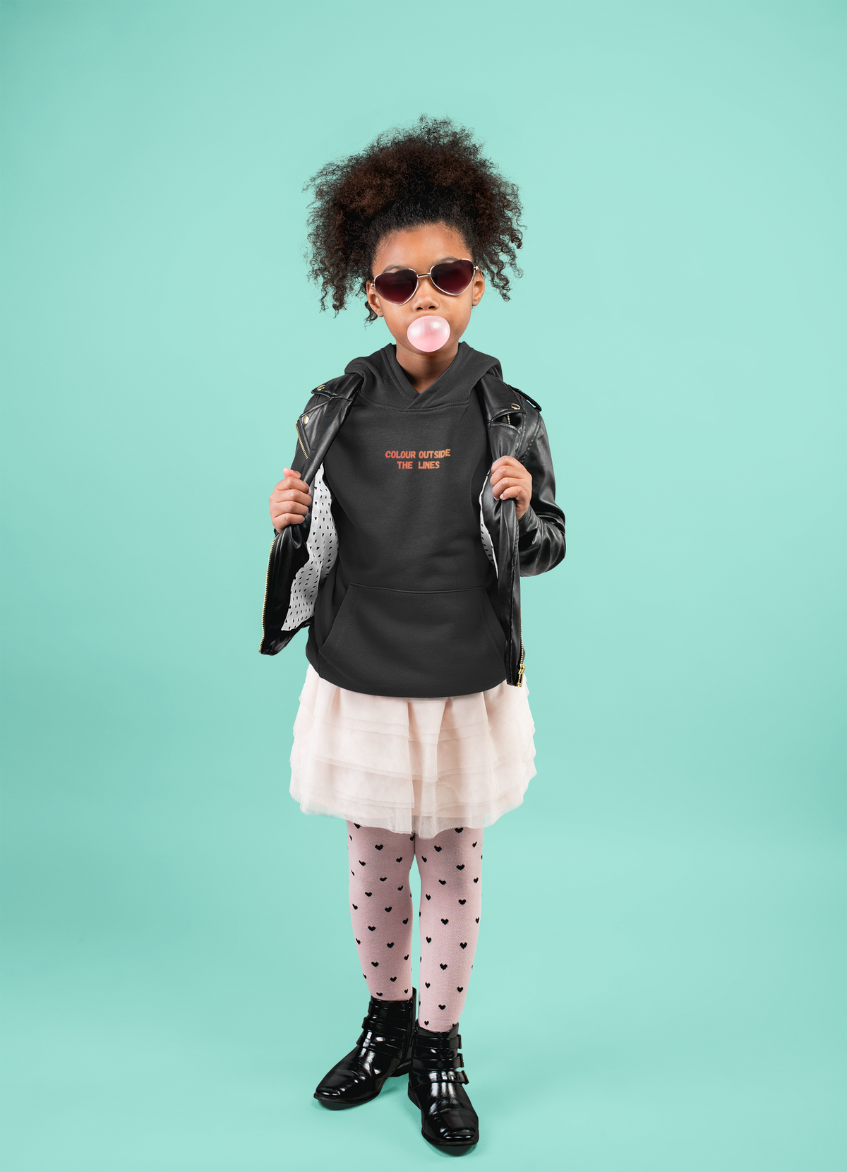 A young girl with wearing a black graphic hoodie, black leather jacket, a light pink tulle skirt, pink heart-patterned tights, and black boots, posing against a solid mint green background.
