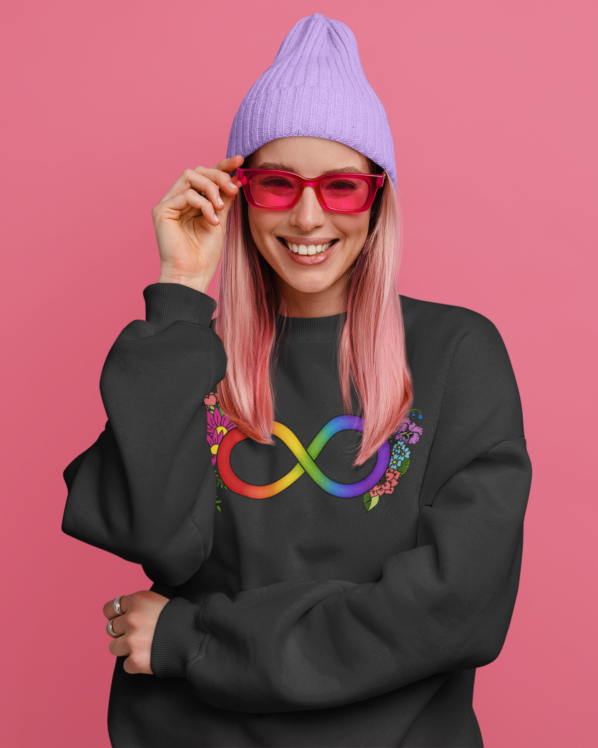 A smiling woman with pink hair wears a lavender beanie, Pink glasses, and a black sweatshirt with a rainbow infinity symbol and floral details on the front, posed against a solid pink background.