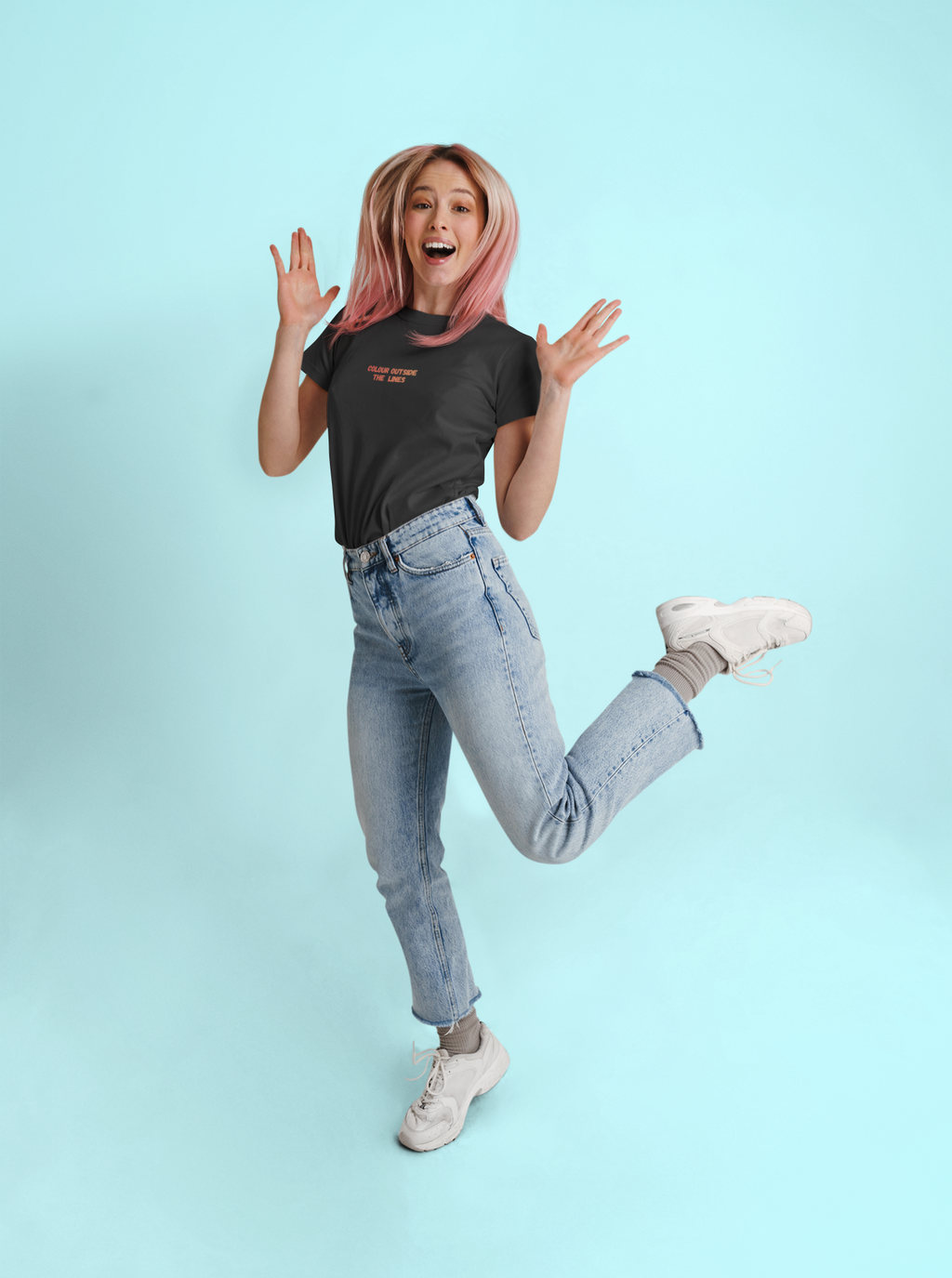 A woman with pink hair jumps in a studio setting against a light blue background, wearing a black graphic t-shirt, light-wash blue jeans, gray socks, and white sneakers.