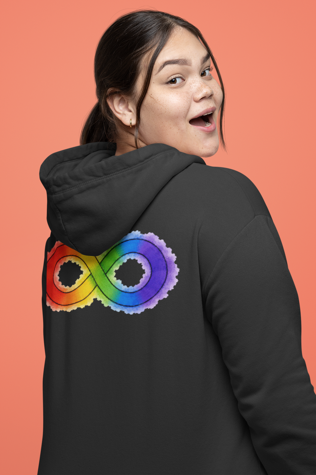 A woman wearing a black hoodie with a large rainbow-coloured infinity symbol printed on the back, shown against a solid coral background.