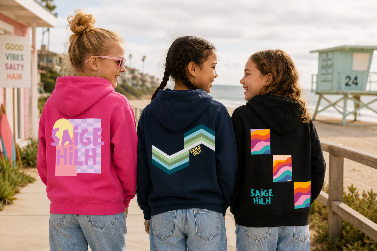 Three girls standing on a beach boardwalk wearing matching SAIGE HILH hoodies in pink, navy, and black, each with a colourful geometric graphic on the back.