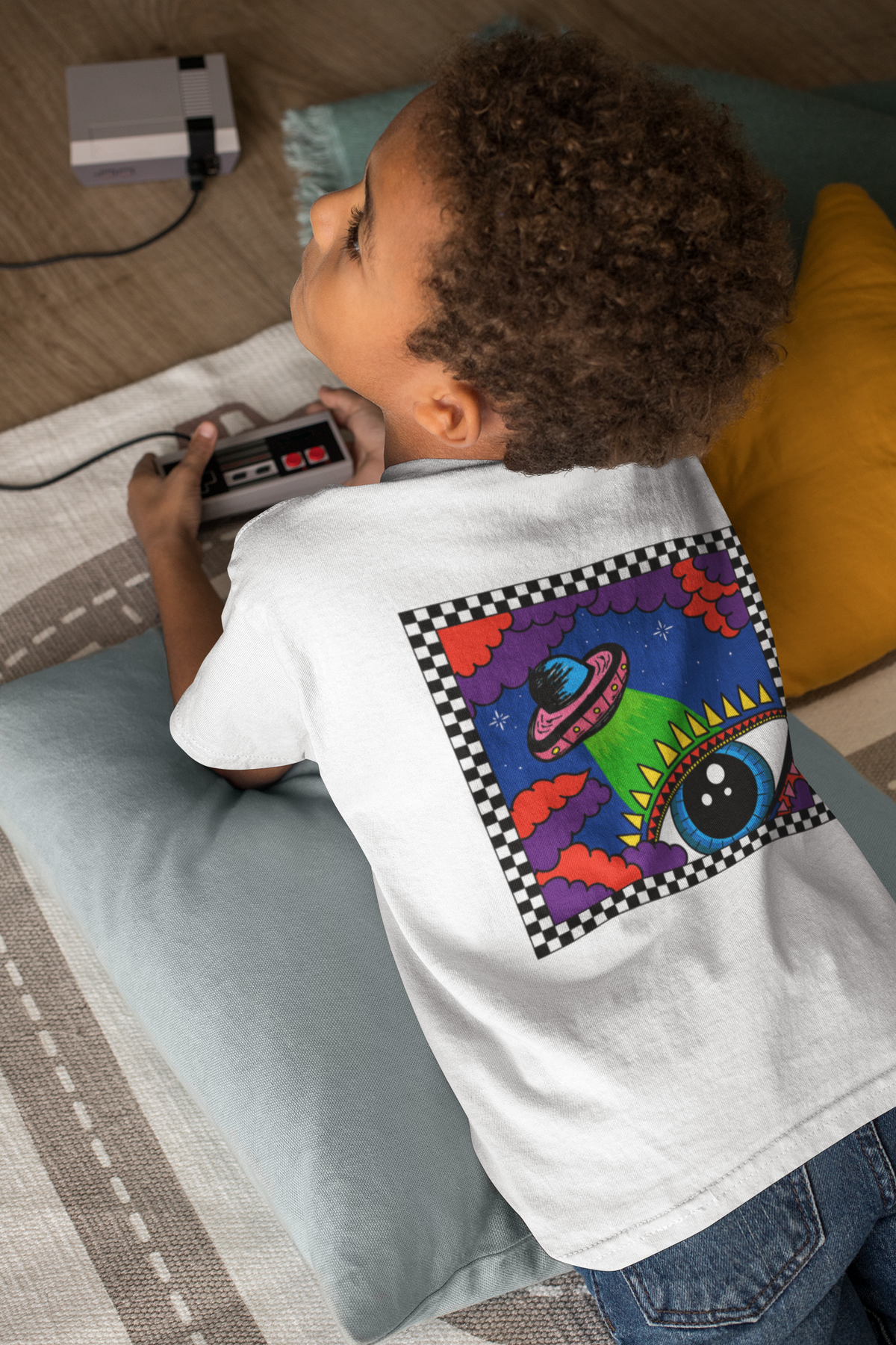 A child lying on a striped floor cushion, wearing a white t-shirt with a large colourful retro alien graphic on the back. The child holds a grey retro game controller, with a vintage Nintendo console visible in the background.