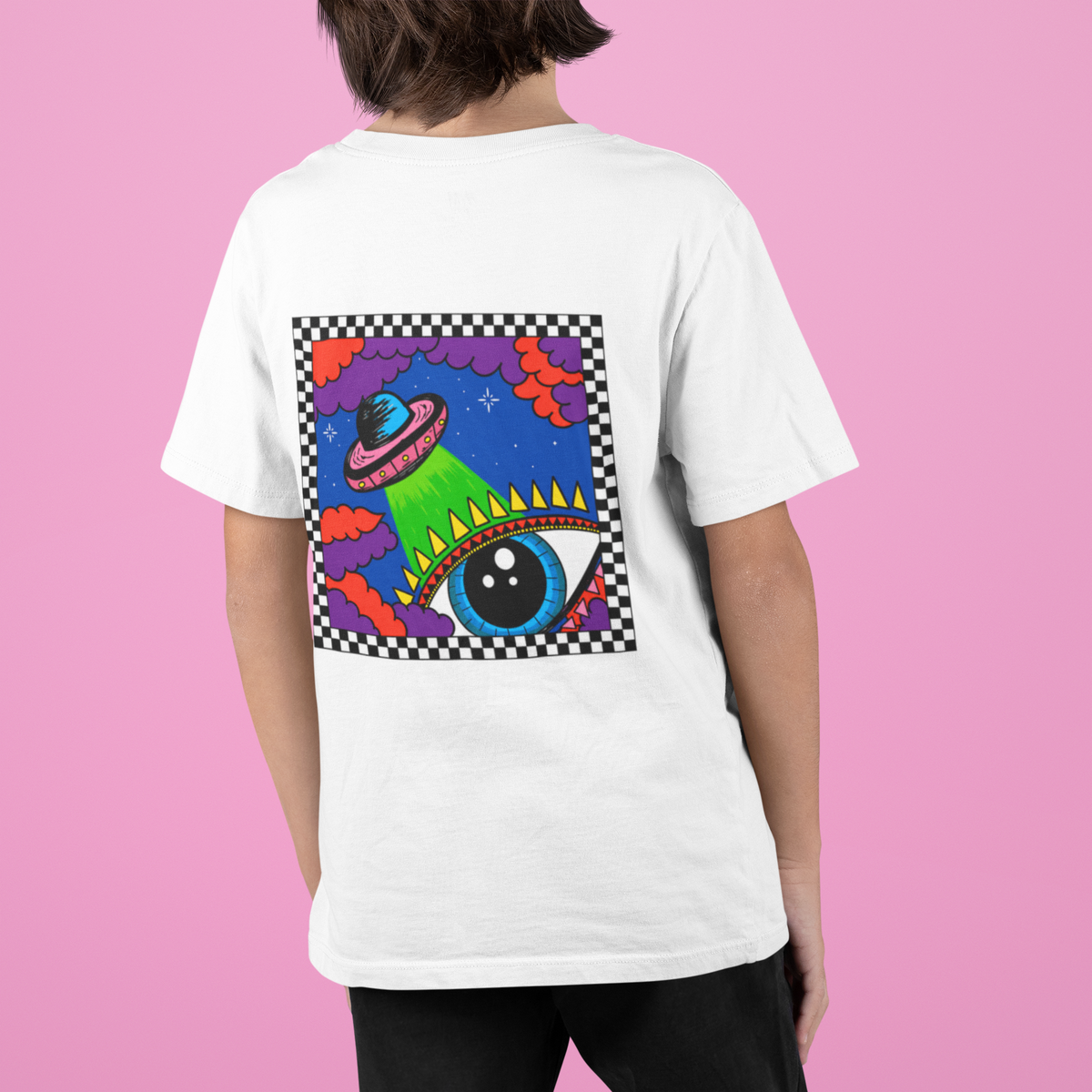 A child wearing a white t-shirt, viewed from the back, featuring a colourful retro sci-fi graphic of a green alien eye and UFO on a checked border. The child is standing against a solid pink background.