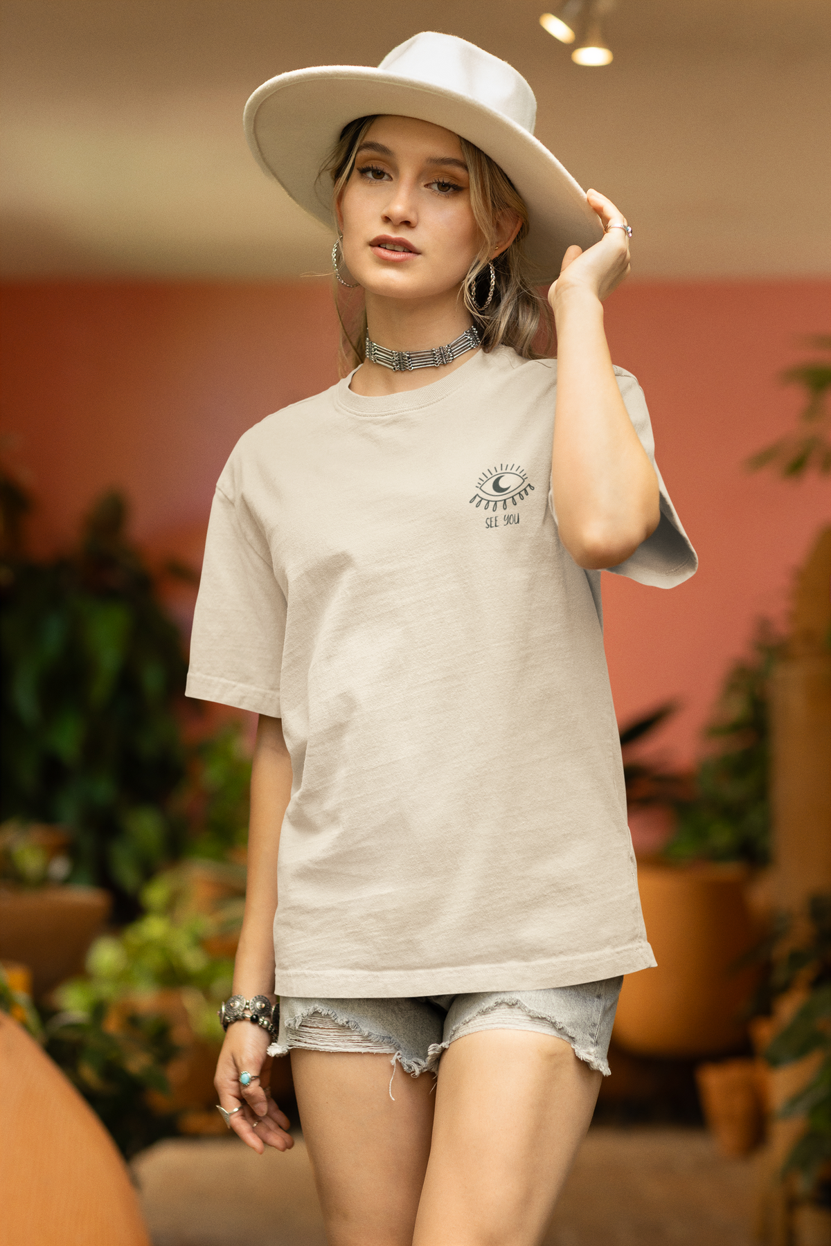 Person wearing a sand-coloured oversized graphic tee featuring a small eye illustration and “See You” text on the left chest.