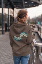 A woman viewed from behind wearing a brown hoodie with a large chevron graphic in shades of teal, green, and white on the back. The graphic features the text 'SAIGE HILH'.