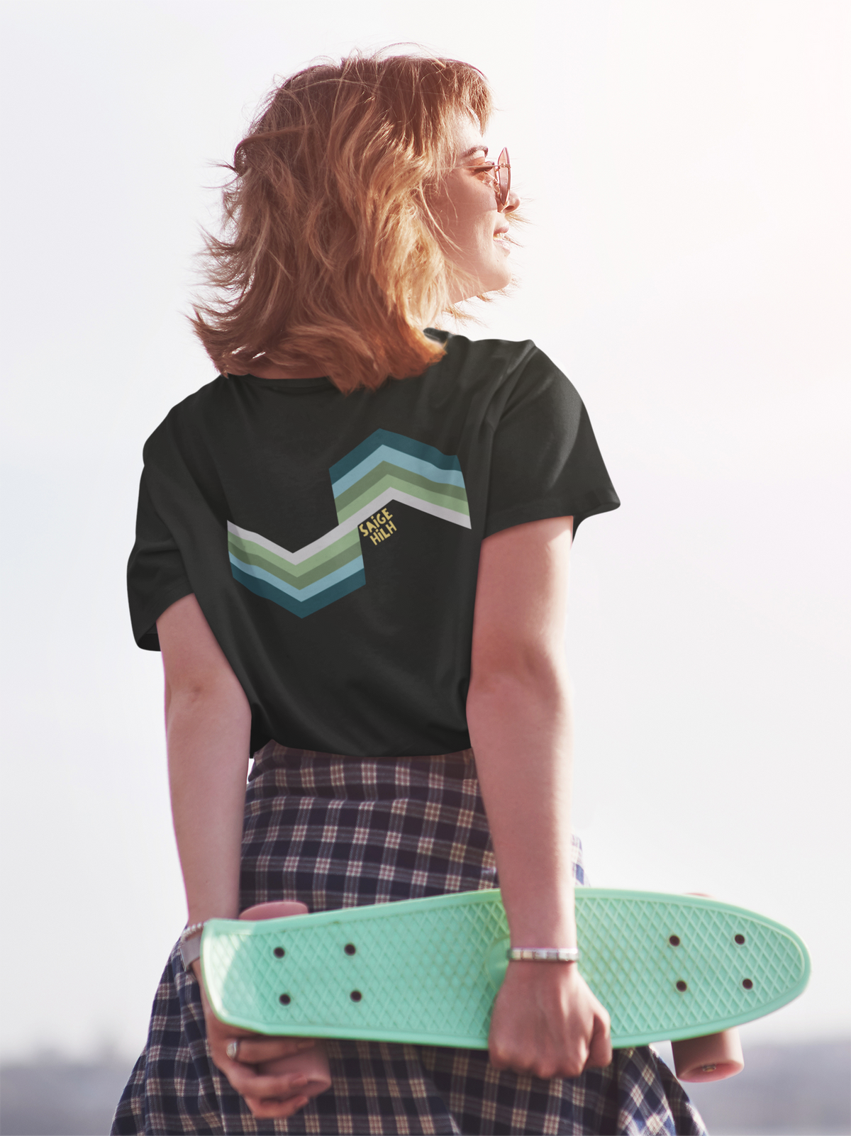 A black short-sleeve crewneck t-shirt with a geometric green and light blue logo on the back, on a female holding a skateboard