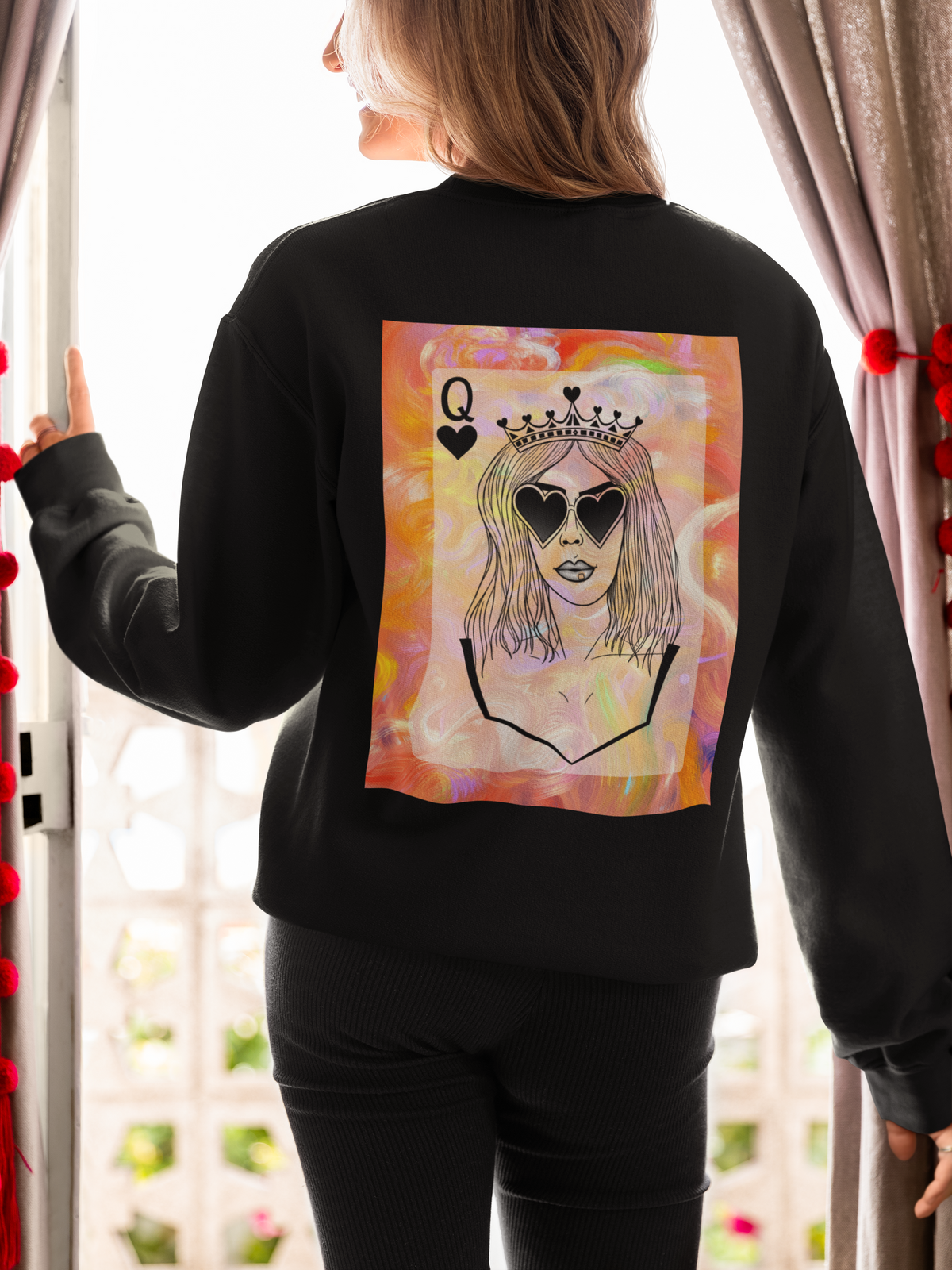 A woman wearing a black sweatshirt with a large back graphic of a queen playing card illustration, shown from behind in a brightly lit room.