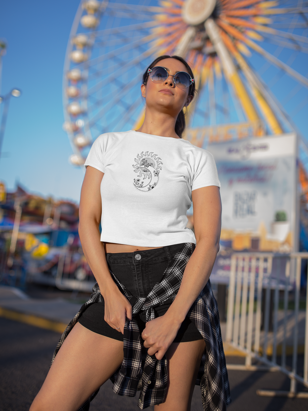 Woman wearing a white t-shirt with text and graphics in front of a Ferris wheel at an amusement park.