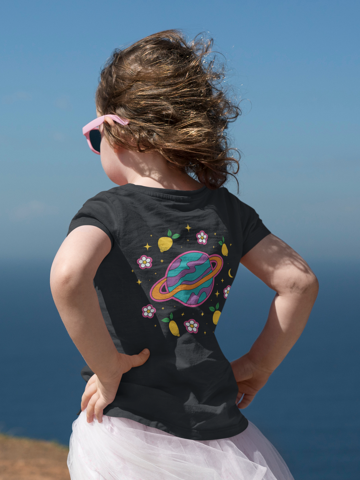 Back view of a child standing outdoors beside a body of water, wearing pink sunglasses, a black graphic T-shirt and a light tulle skirt. The colourful back design features a purple and teal ringed planet surrounded by lemons, flowers, stars and a crescent moon against a bright blue sky background.