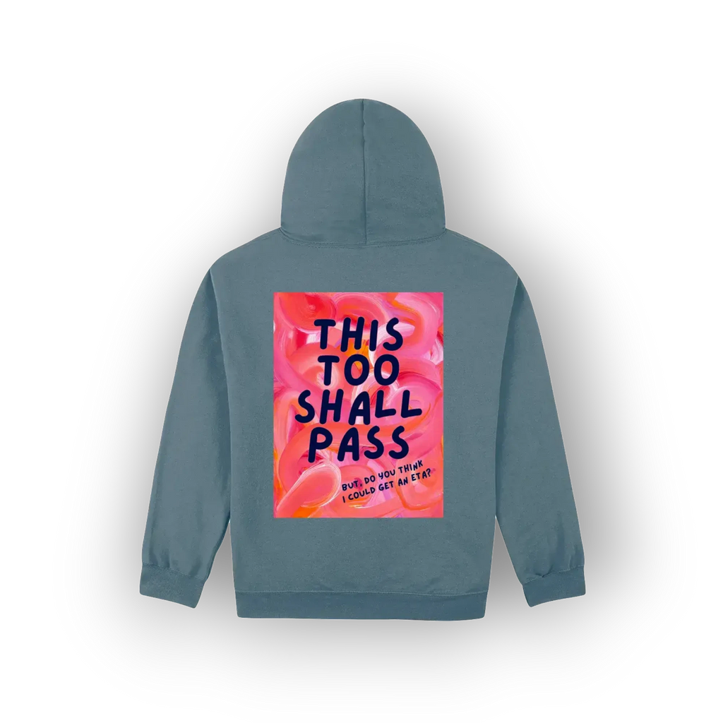 A slate blue hoodie shown from the back, featuring a large square graphic print with a pink and orange floral background and the text 'THIS TOO SHALL PASS' in dark blue lettering.