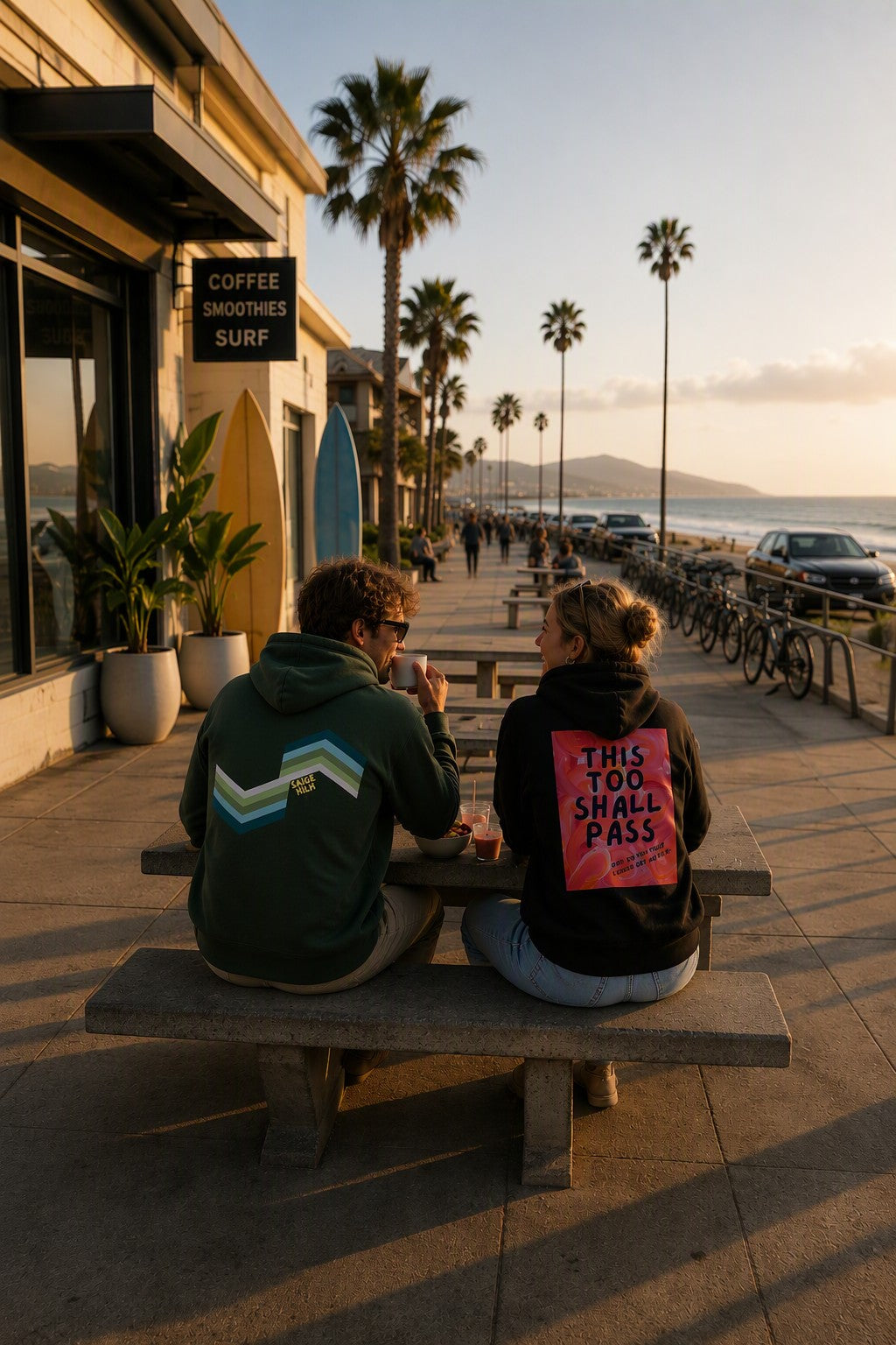 Two people sitting on a concrete bench at a seaside boardwalk at sunset, viewed from behind. One wears a dark green hoodie with a light blue and white chevron design on the back, and the other wears a black hoodie with a bright pink graphic on the back that reads 'THIS TOO SHALL PASS'.