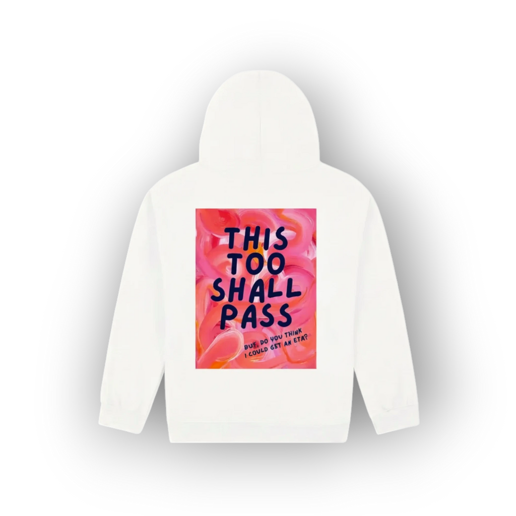 A white hooded sweatshirt shown from the back, featuring a large square graphic print with a pink and orange floral background and the text 'THIS TOO SHALL PASS' in dark blue lettering.