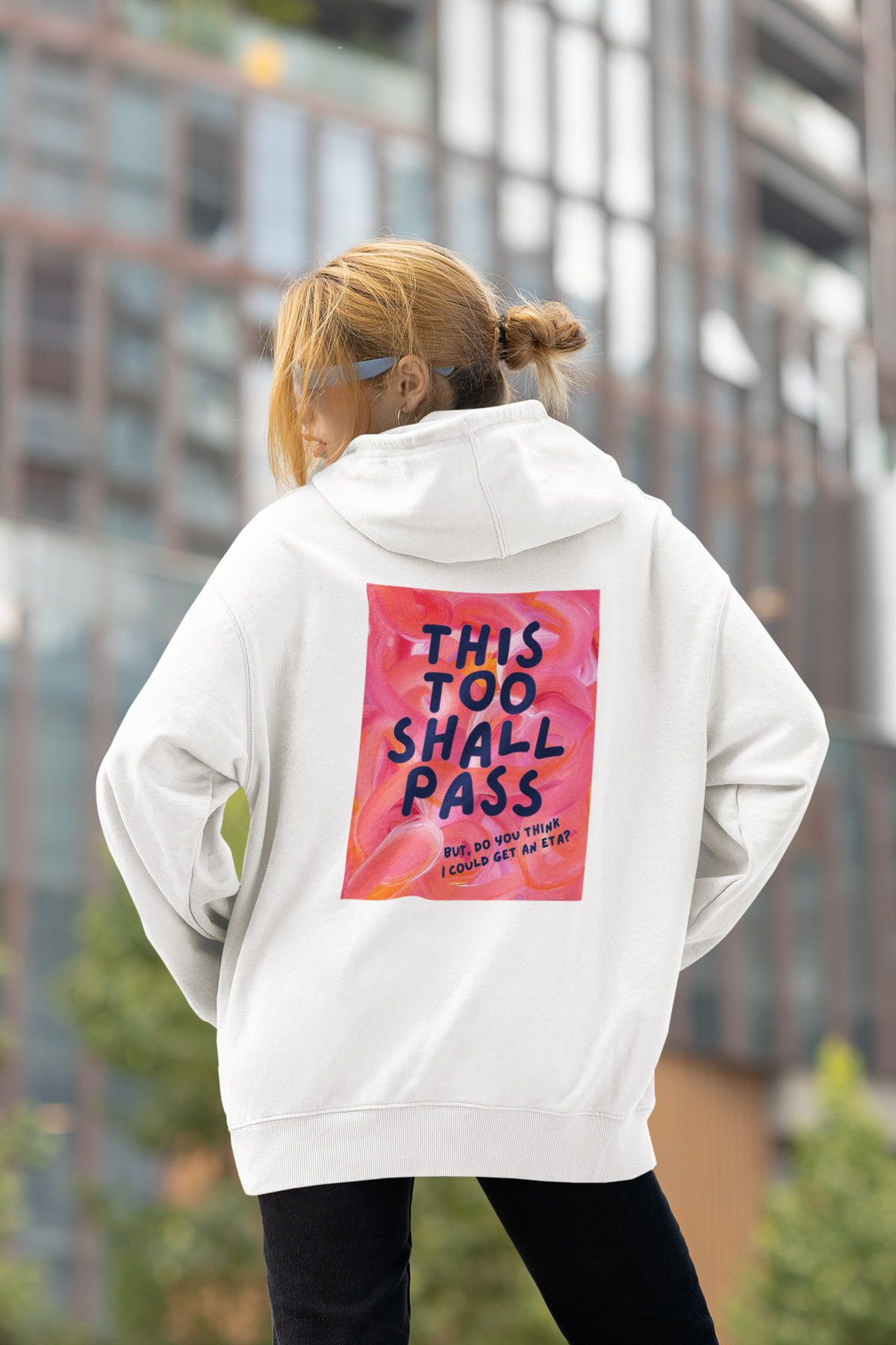 A white hoodie shown from the back, on a female model featuring a large square graphic print with a pink and orange f background and the text 'THIS TOO SHALL PASS' in dark blue lettering.